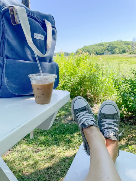Girl wearing Converse shoes with feet up on bench. Backpack and coffee on the table.