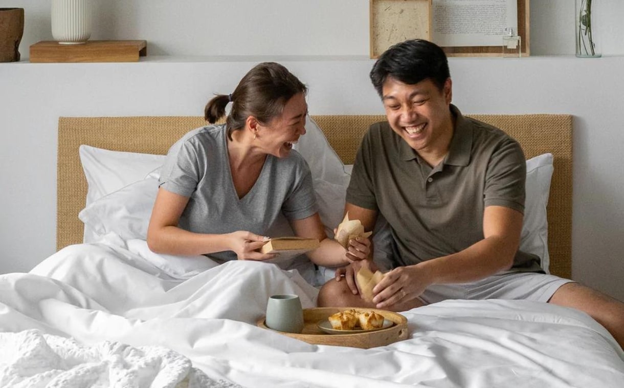 man and woman laughing while sitting on bed with white sheets