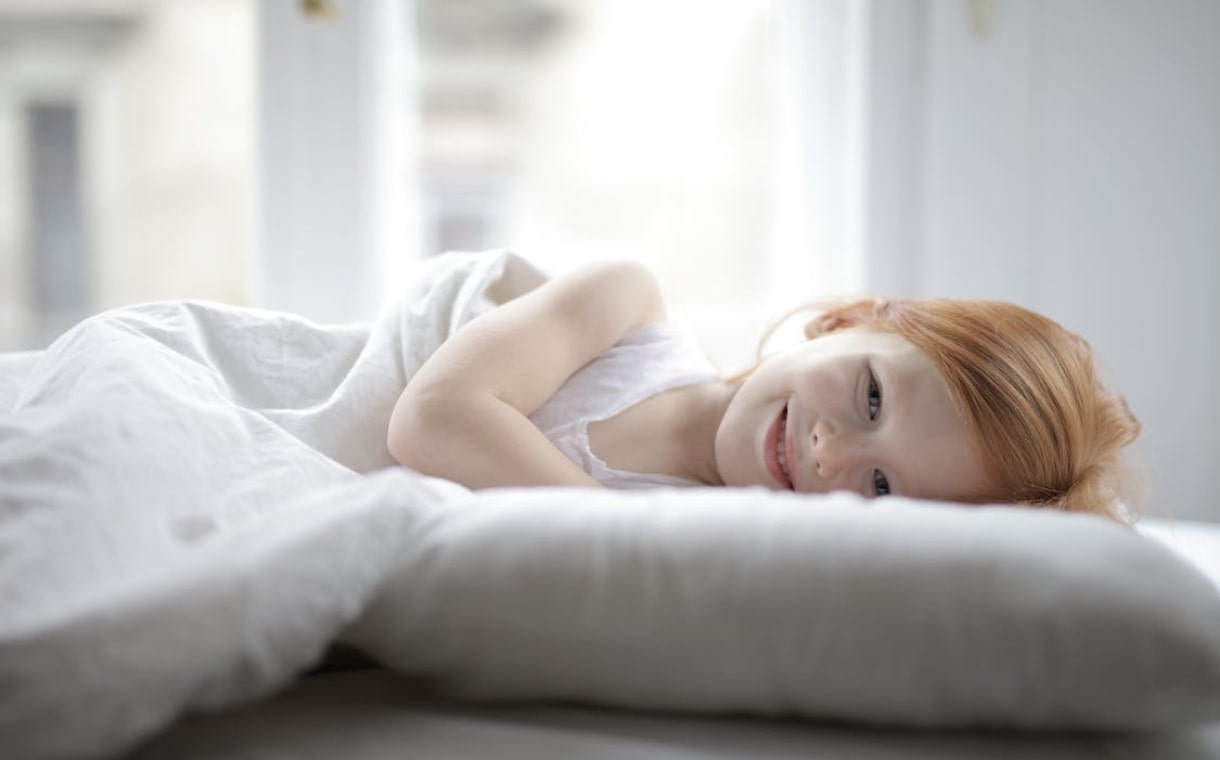 little girl lying on white bed while smiling at the camera