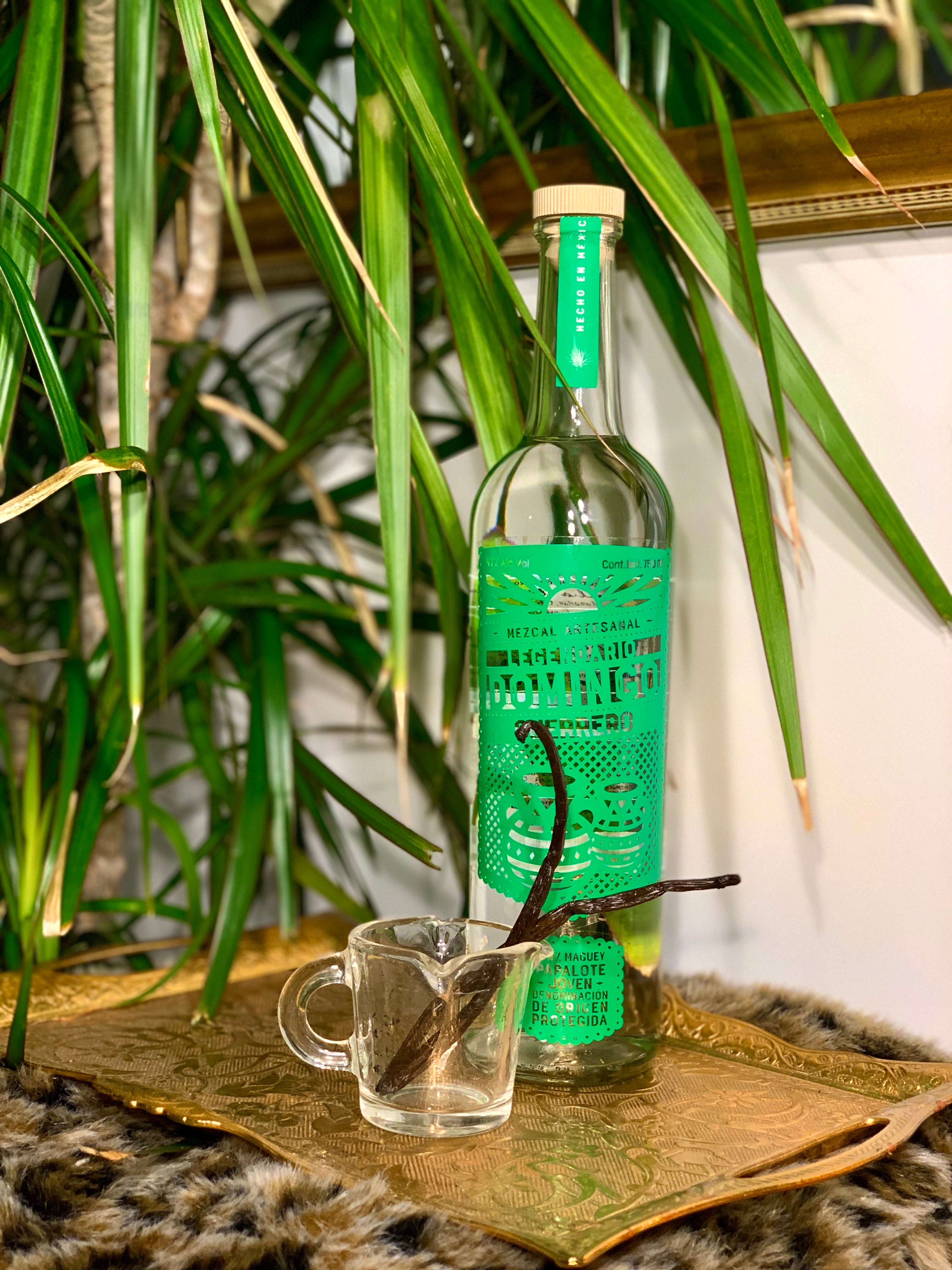 Photo of coffee shot glass with two vanilla beans in it and mezcal bottle with green accents, on a golden tray