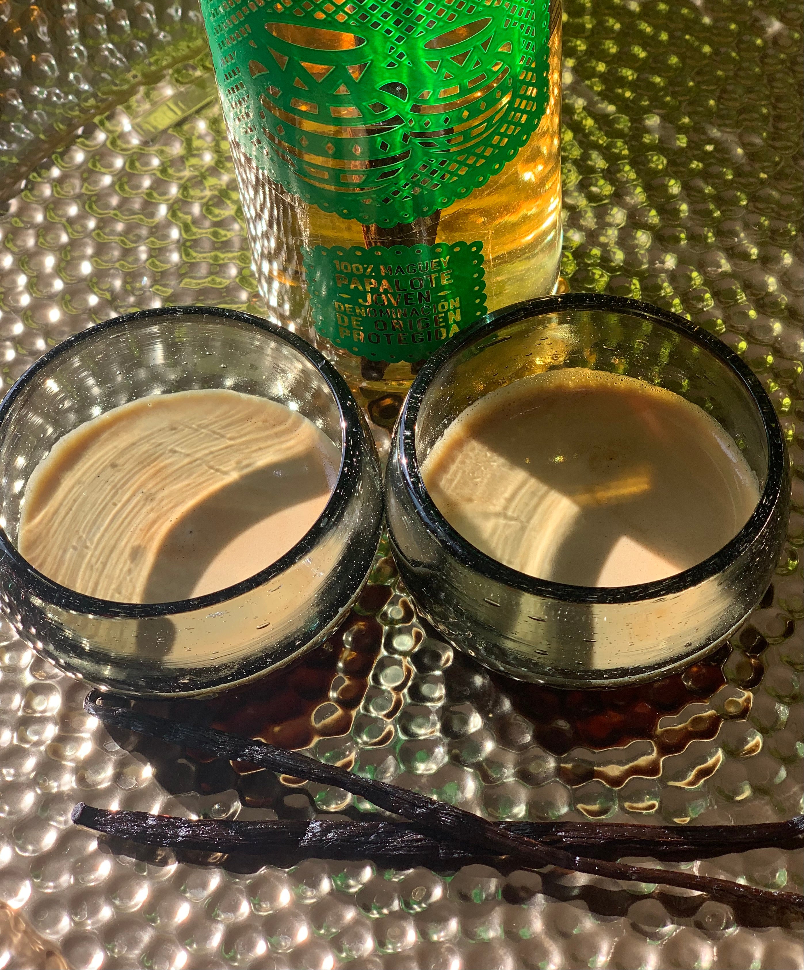two capitals made of black glass filled with espresso, two vanilla beans in front and mezcal bottle in back, all on golden tray