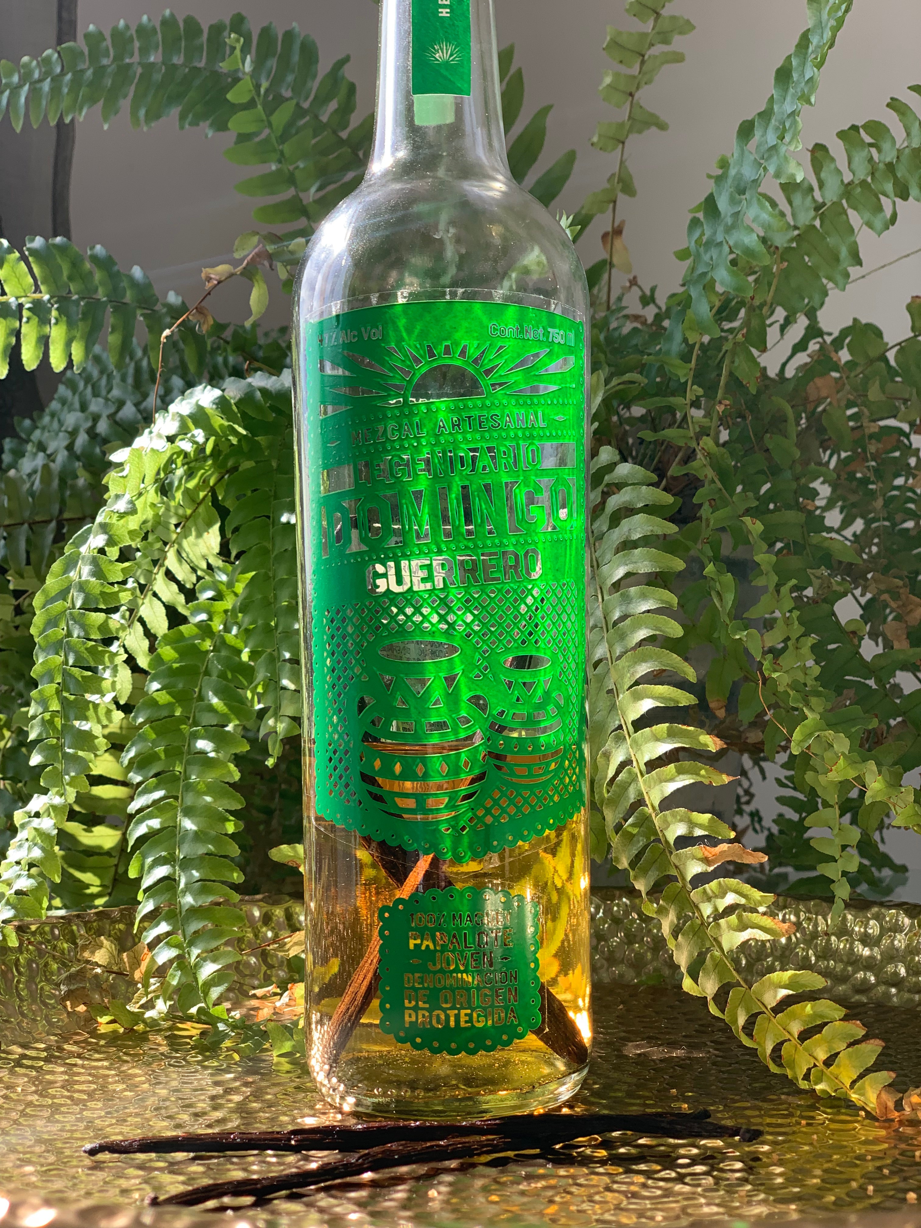 Mezcal bottle with green accents, two vanilla beans in front of it, all on a golden tray
