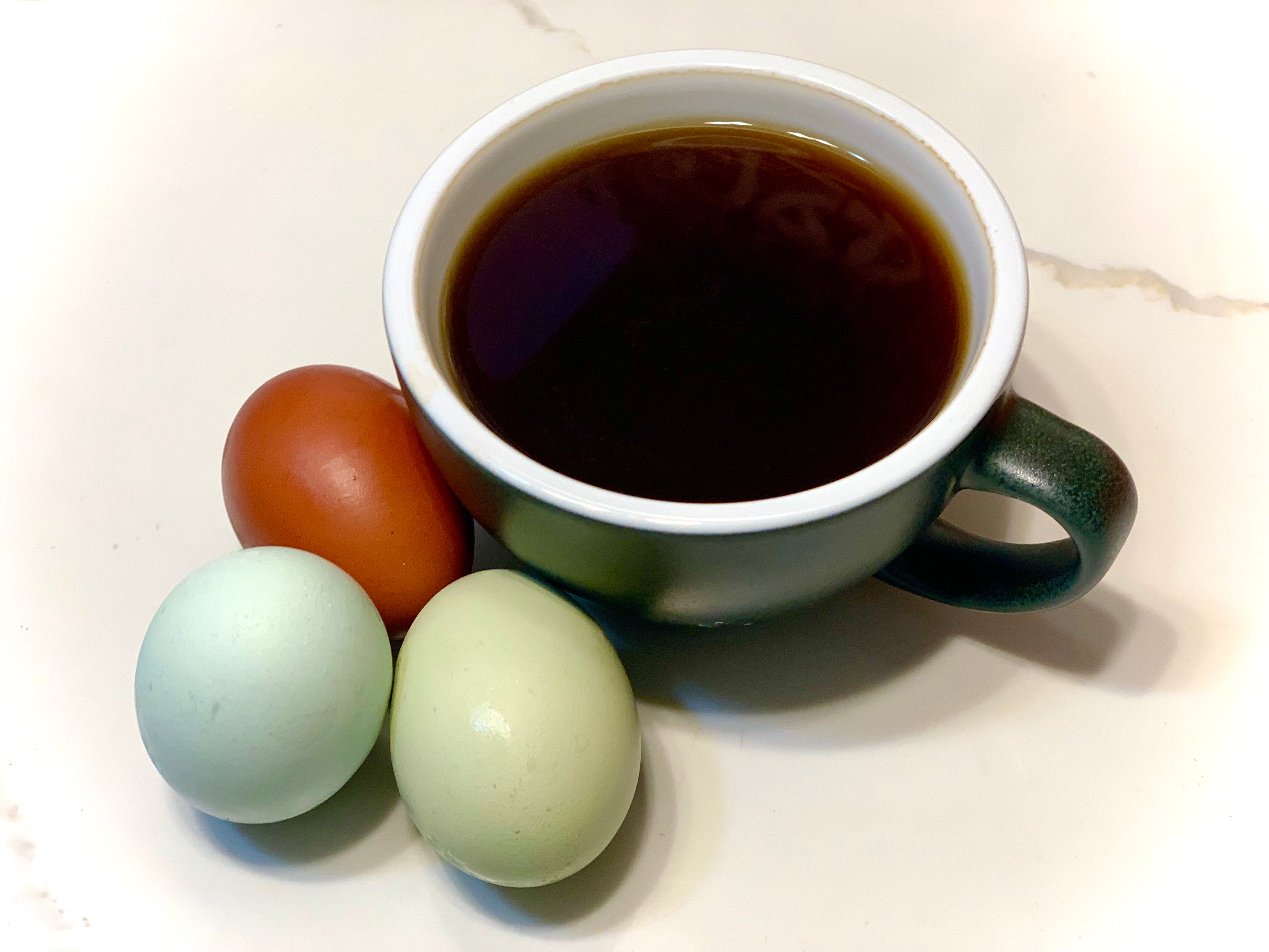 Photo of ceramic coffee cup with coffee, surrounded by four colorful eggs and coffee beans