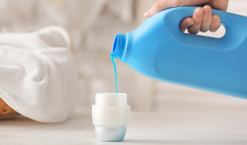 Person pouring a blue bottle of laundry detergent into a white bottle cap