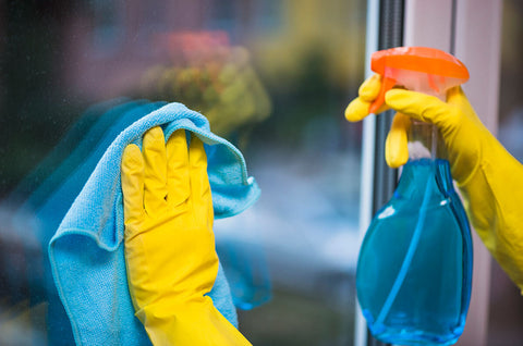 Yellow gloved hands holding spray bottle containing blue liquid and using blue cloth to wipe window