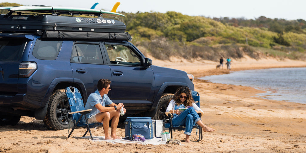 Young couple sitting in backpack chairs at the beach with an SUV behind them with surfboards on top