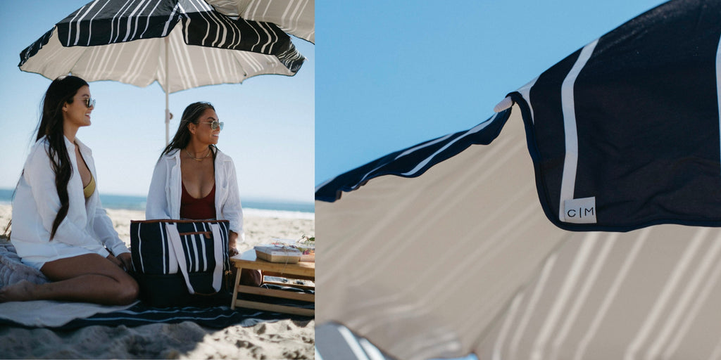 Two young ladies sitting under a navy and white striped umbrella on the beach, enjoying a charcuterie board