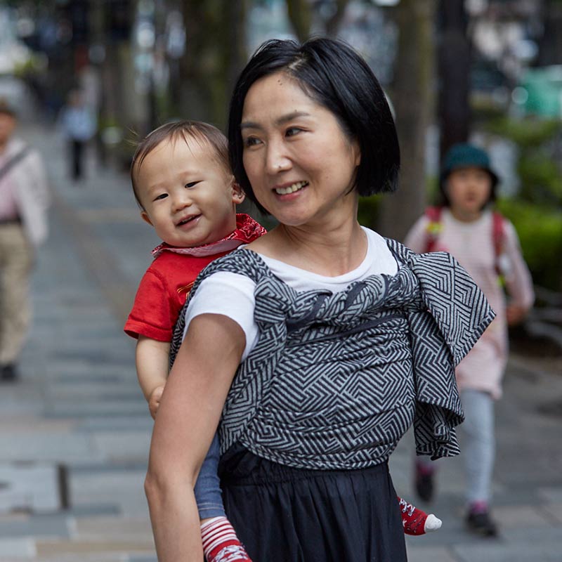 モノクロのベビーラップで１歳の男の子をおんぶしながら表参道を散歩するお母さん