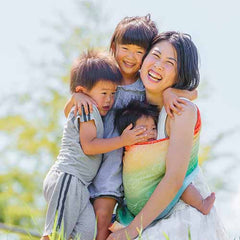 下の赤ちゃんをベビーラップで抱っこしながら、上の二人の兄弟に囲まれて、幸せそうに笑っているお母さん