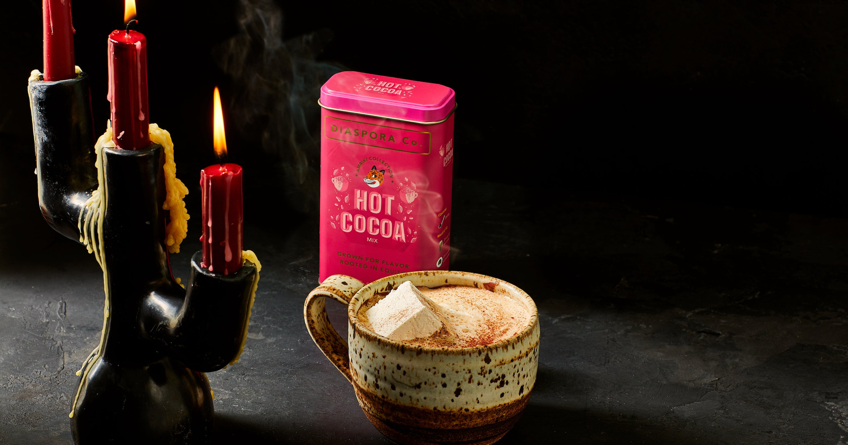 A pink Hot Cocoa tin sits in a line of lite beside a steaming mug of hot cocoa and a lit candleholder