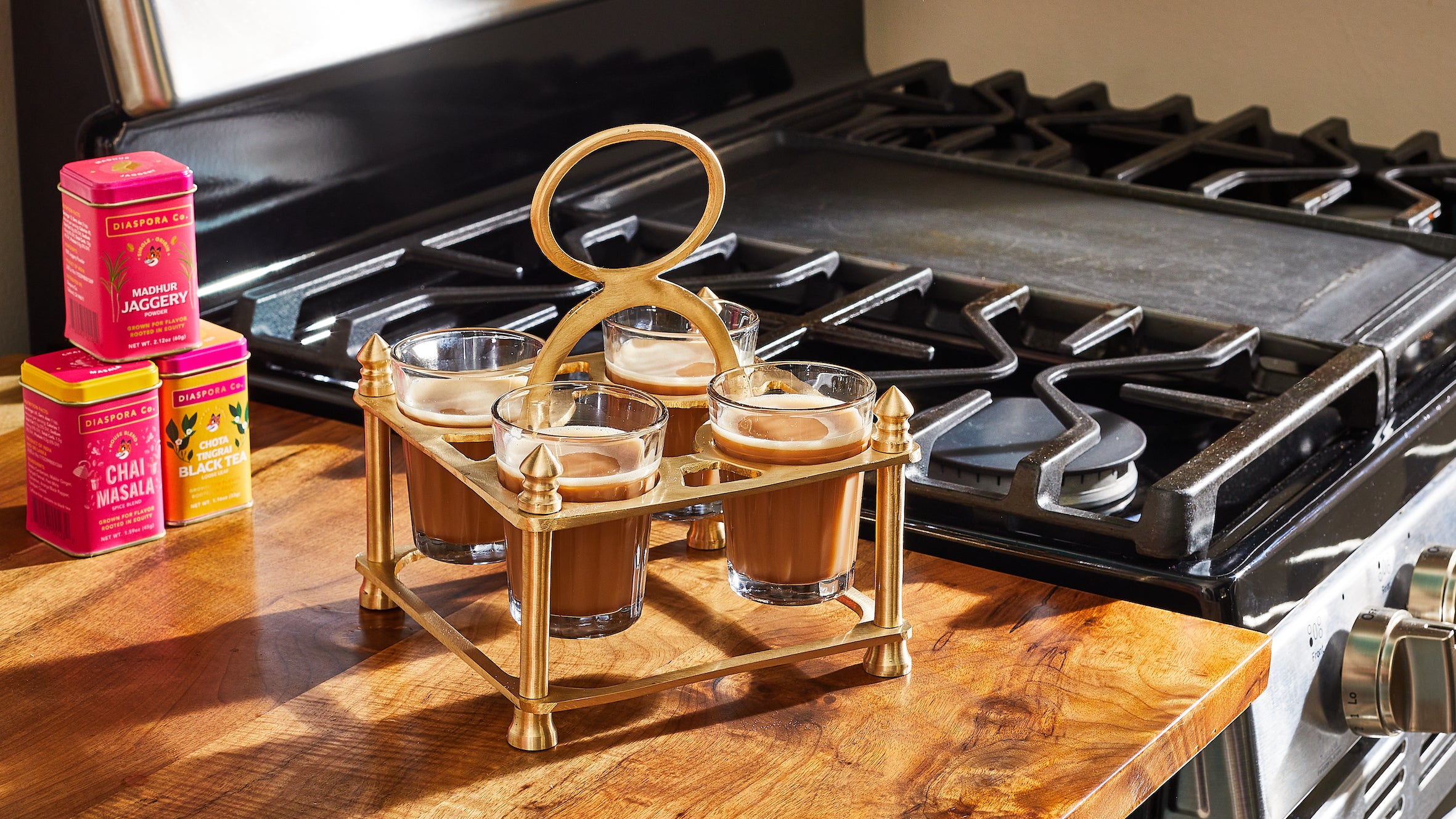 Chai Caddy with 4 cups of chai sitting on a wooden kitchen counter in sunlight. There is a stack of 3 tins in the background of Jaggery, Chai Masala, & Chota Tingrai Black Tea