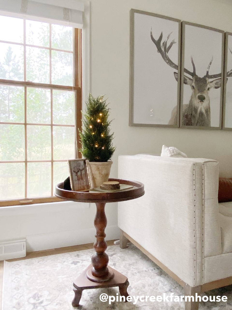 Artificial cedar trees decorated on a tall round table next to a large window and white sofa