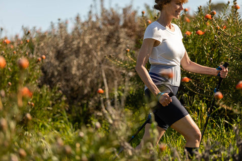 Woman hiking through the bush. She is holding hiking sticks and wearing Bauerfeind's LumboTrain back brace, the benefits of which include improving your posture 