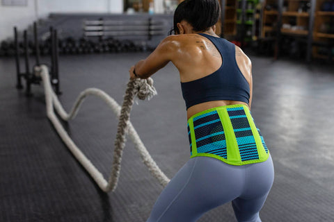Woman flicking battle ropes at the gym. The shot focuses on her torso and upper body. She is wearing Bauerfeind's Sports Back Support brace to help protect her lumbar from injury 