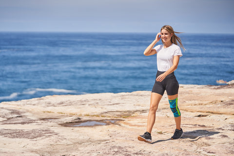 Woman walking along a beach in running shoes and Bauerfeind's Sports Knee Support to protect her knee