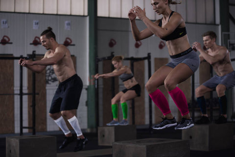 Men and women in cross fit class doing box jumps as part of their leg day routines. They are wearing Bauerfeind's Compression Calf sleeves in various colours.