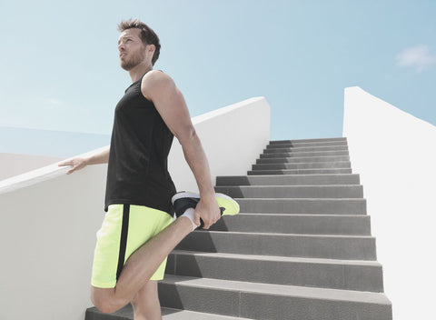 Man in yellow shorts standing on a stairwell. He is doing the quad stretch (holding his left heel to his glutes), which is good for relieving tension in the quadriceps after leg day.