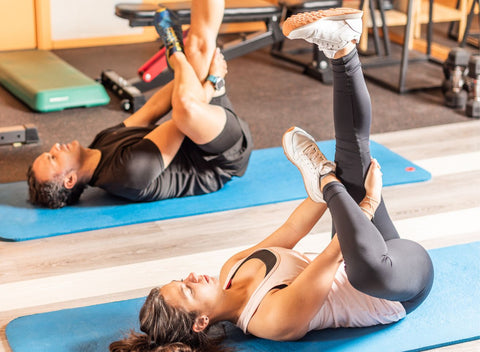 Man and woman stretching in an exercise studio. They are laying on their backs on their yoga mats with their right legs bent and left legs straight in a figure 4 position.