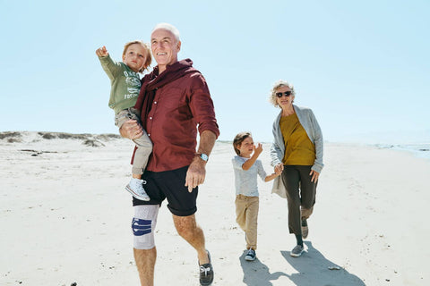 Older man walking in the beach with his family. He is holding his grandchild and smiling. He's wearing Bauerfeind GenuTrain A3 brace, a great knee brace for mild Osteoarthritis.