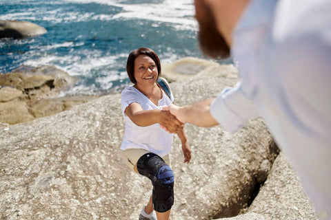 Woman at the beach getting help from a man to climb up a rock. She is wearing Bauerfeind's GenuTrain OA Knee Brace to manage knee arthritis, which can cause knee pain without injury