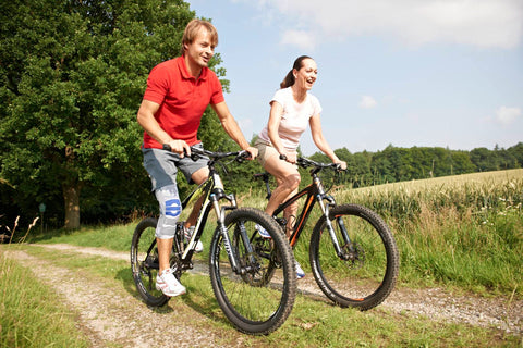 Man and woman cycling through the park on a sunny day. The man is wearing Bauerfeind's SofTec Genu Knee Brace to assist in his recovery from injury and reduce re-injury risk
