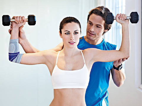 Woman exercising in the gym wearing Bauerfeind elbow support, man advising her on the exercises. 