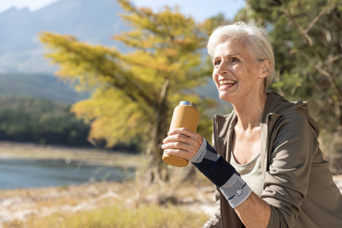 Older woman with a sprained wrist sitting in a park and drinking water. She is wearing Bauerfeind's ManuTrain wrist brace.