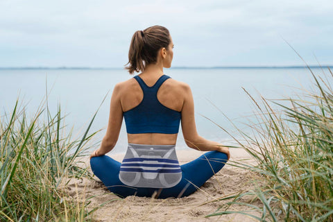 Woman sitting cross-legged at the beach in athletic wear. She is wearing Bauerfeind's LumboTrain Back Brace, a great brace for relieving pain in the lower back