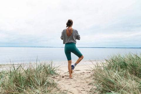 Woman doing the Tree Pose on the beach on a chilly day. She is wearing a long sleeve top, leggings, and a Bauerfeind ankle brace for her joint pain