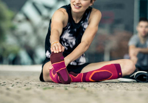 Woman sitting on the floor and stretching her hamstrings. She's wearing pink Bauerfeind Performance Socks, which have padding around the toes for protection