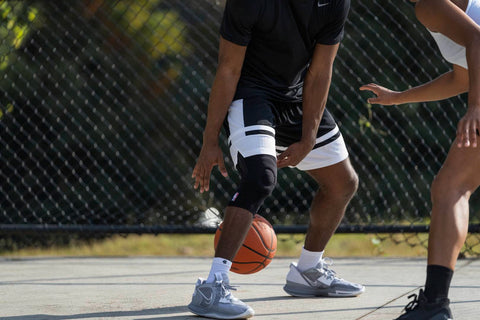 Man dribbling a basketball between his legs. He is wearing basketball gear consisting of loose gym clothes, basketball shoes, and Bauerfeind's NBA Sports Knee Support