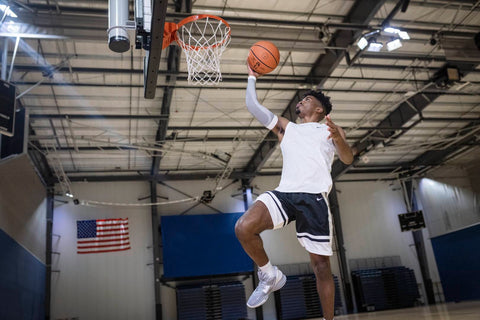 Man playing basketball in the winter in an indoor court. He's jumping up to make a slam dunk and wearing Bauerfeind's Sports Compression Arm Sleeve.