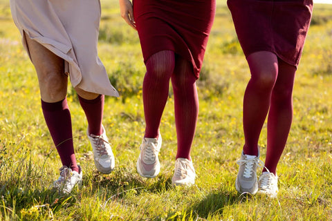 3 women walking through a grassy field wearing Bauerfeind's VenoTrain stockings of various lengths: knee-high, thigh-high, and pantyhose, all in maroon.