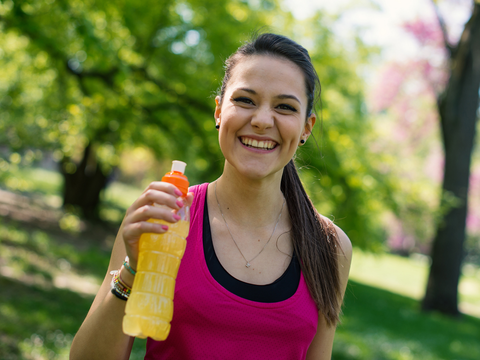 Woman at the park taking a break from running. She is smiling while holding up a bottle of sports drink