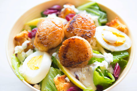 A Mediterranean seafood dish containing grilled scallops, eggs, leafy greens, and cooked fish, which are believed to be good for managing osteo and rehumatoid arthritis