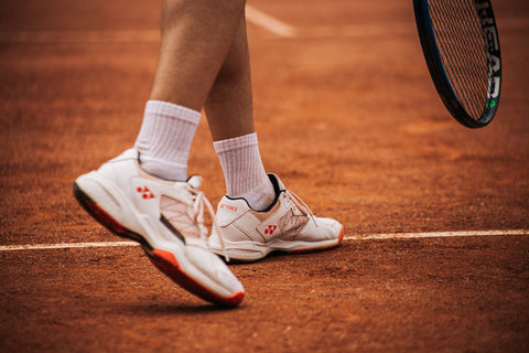 Person wearing well-fitted white tennis sneakers, good shoes for avoiding tennis injuries like tennis toe. The shot focuses on the shoes, orange court floor, and a bit of their racquet.