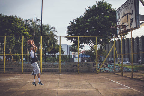 Wide shot of a man playing basketball. He is in the middle of making a shot, jumping up with the ball just about to leave his hands. He is wearing sneakers to achieve good foot support while he plays 