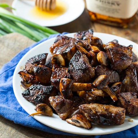 Image of finished steak bites and mushrooms plated and sitting on a table in front of a bottle of bourbon and small dish of honey.