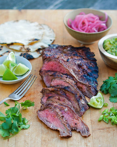 Image of fully-cooked flat iron steak with half of the steak already carved on a cutting board. Side dishes are purple onions, lime wedges, fresh cilantro, and homemade tortillas.