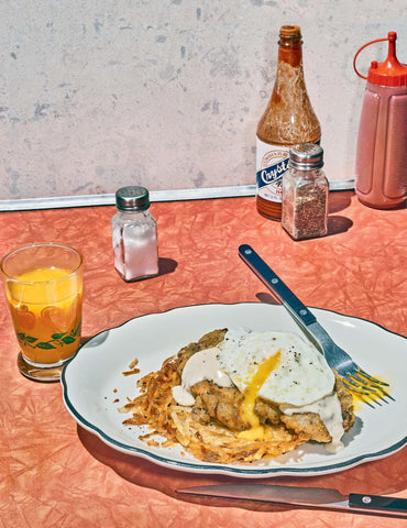 Image of plated chicken fried steak meal sitting on a counter with a fork and knife, glass of orange juice, salt and pepper shakers, bottle of hot sauce, and a bottle of ketchup,