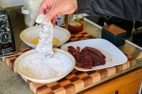 Image of human hand holding a piece of steak that has been dredged through the beaten and the flour.