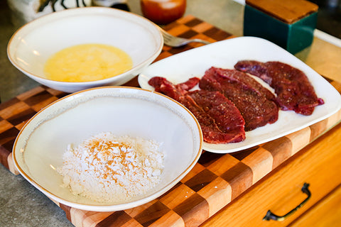 Image of elk dredging station with a bowl of beaten egg, a bowl of flour, and the plated raw steaks.