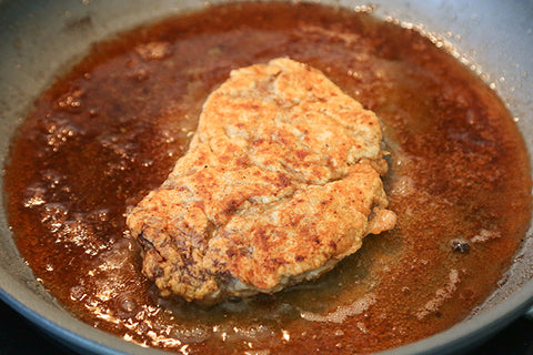 Image of a piece of the elk steak frying in a frying pan atop a stove.