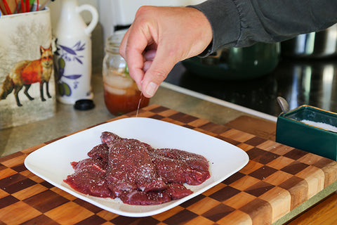 Image of human hand salting serving size pieces of raw elk meat.