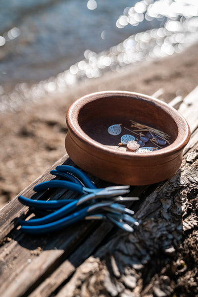 A clay bowl laying on sand with different pieces of jewelry inside it, next to a pile of blue pliers.