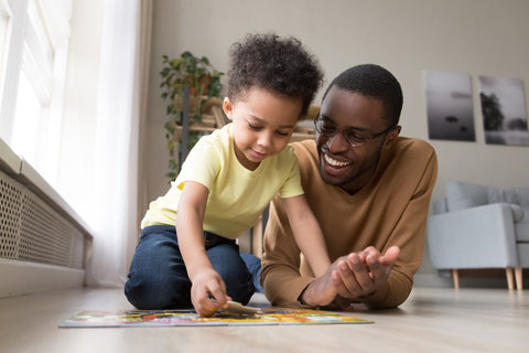 An African American father and his toddler son assemble a floor puzzle on the floor of their home. Minimal, modern, yet warm furnishings are visible in the background of the image.