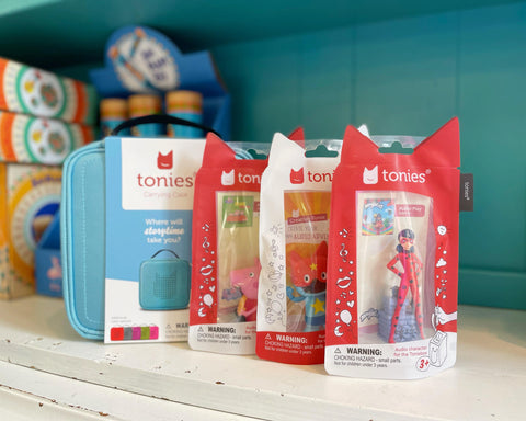 three different Tonies lined up on a white shelf against a turquoise blue background, with blue Tonies headphones in the lineup as well