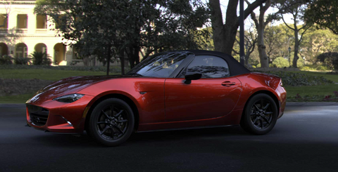 A car with red and black combination which is a Mazda MX-5 Miata sitting at a road with green grass and trees