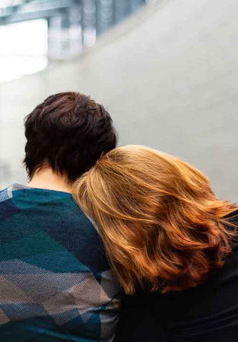 Two people sitting next to each other, offering comfort. one with head on other person's shoulder