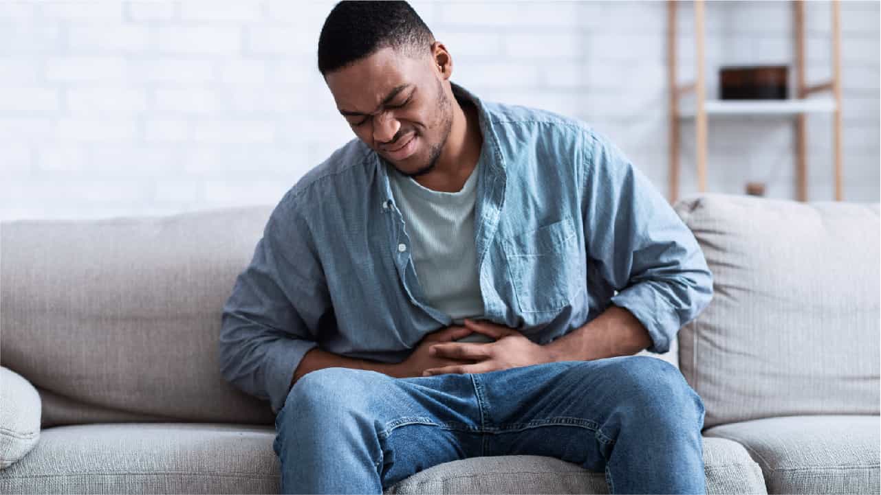 How Much Vitamin C Do You Need? Determine your upper limit with a test where high-dose vitamin C on an empty stomach is taken until gastric distress, pictured as a man sitting on a couch and grimacing as he holds both hands over his abdomen in discomfort